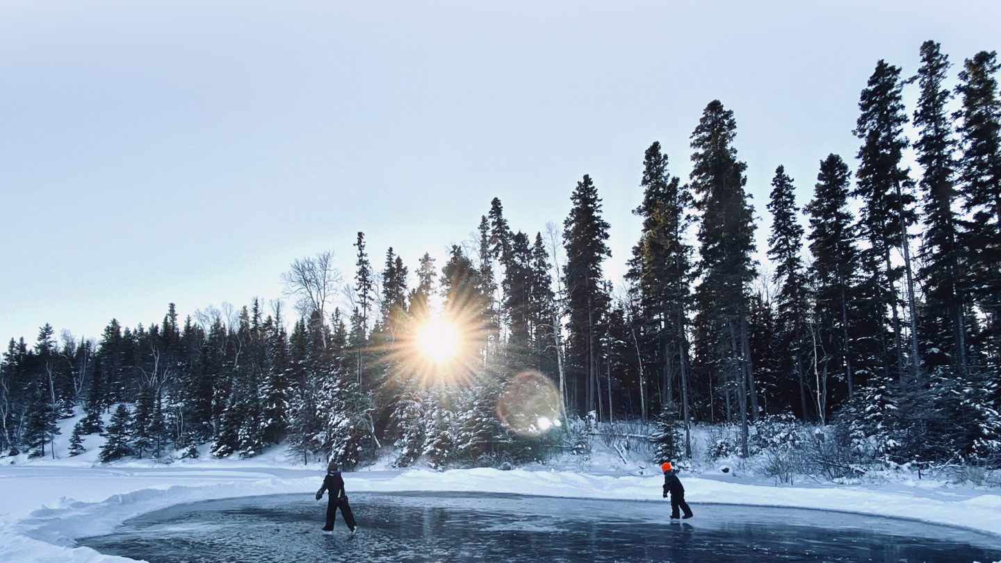 Two kids skating on a pond on a sunny winter day amongst tall pine trees